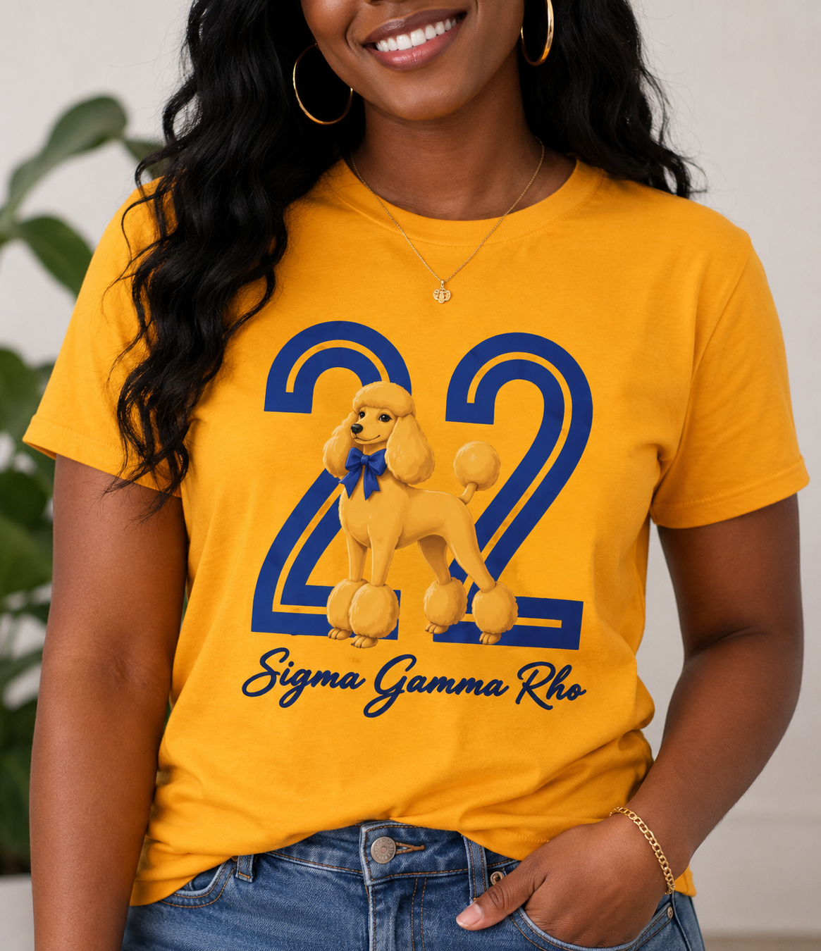 
Smiling woman wearing a gold Sigma Gamma Rho sorority-inspired T-shirt featuring a large blue number 22 with a golden poodle wearing a blue bow centered on the design. The words “Sigma Gamma Rho” appear in blue script beneath the graphic. The model is shown from the shoulders to the hips, wearing jeans, gold hoop earrings, and a delicate necklace.